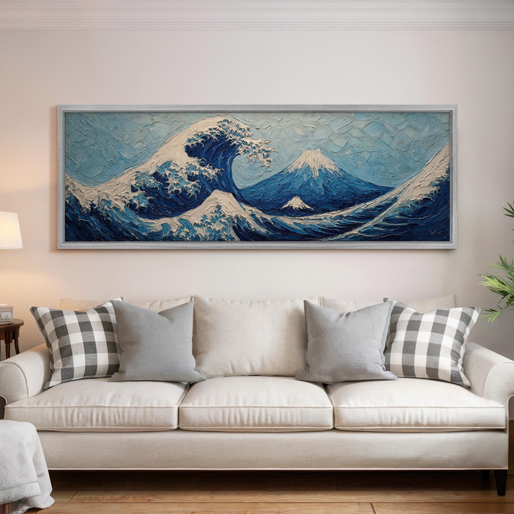 a large, framed painting of a wave with mountains in the background, hanging on a wall above a white couch with gray and white plaid throw pillows.