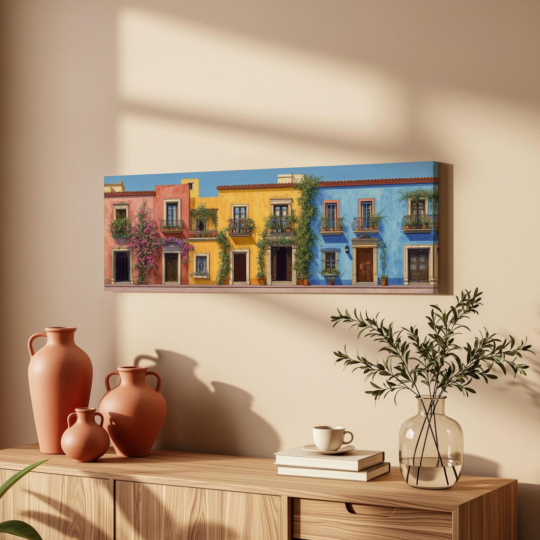 a wooden dresser with a vase of greenery, a stack of books, and a ceramic vase with flowers. Above the dresser, there is a painting of colorful buildings with balconies and a blue wall.