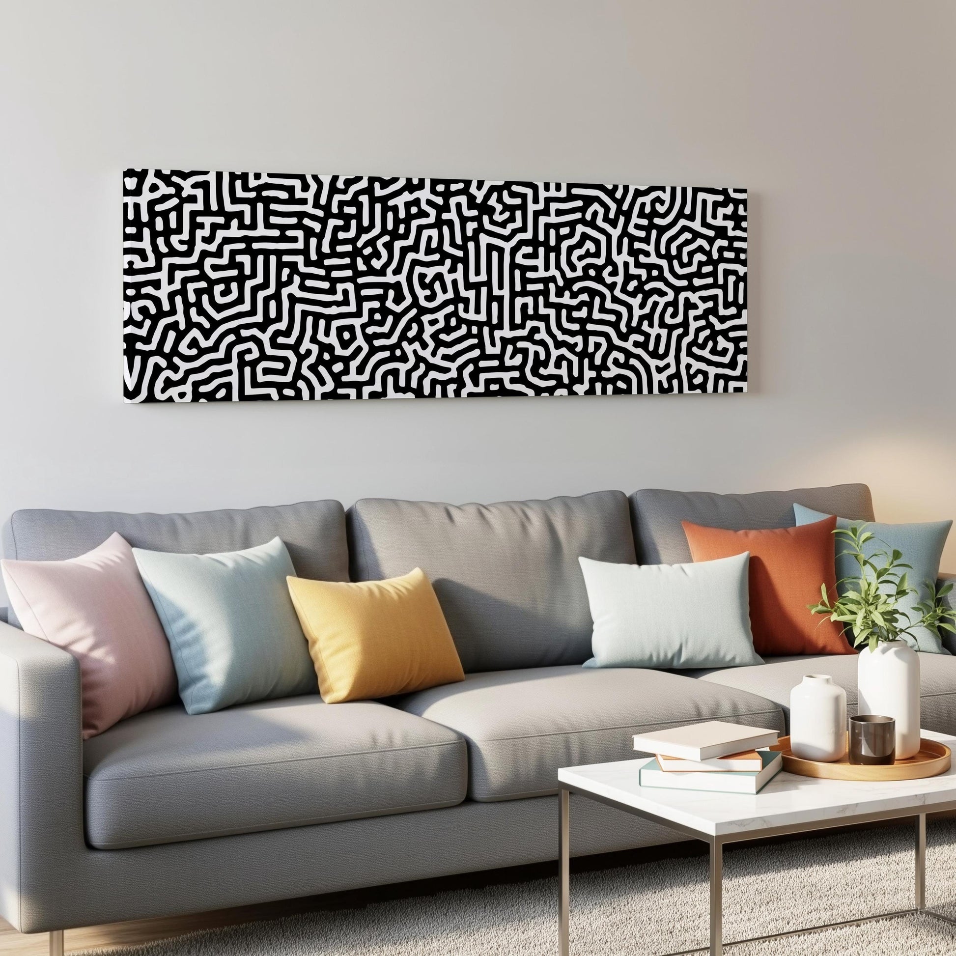 a modern living room with a gray sofa adorned with colorful throw pillows. Above the sofa, there is a large abstract artwork with a black and white pattern. The room also includes a coffee table with books and a plant, creating a cozy and inviting atmosphere.