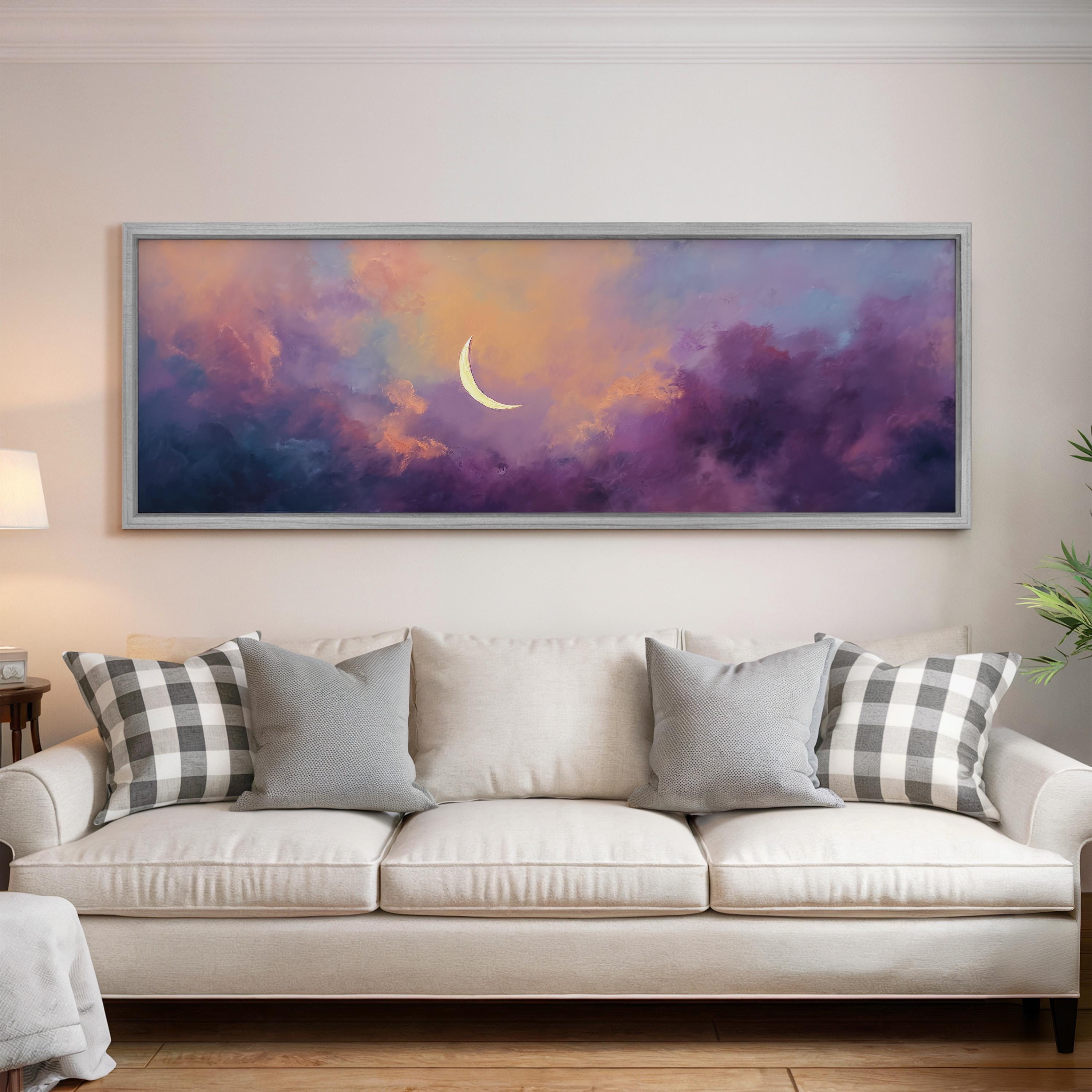 a large, framed painting of a crescent moon set against a colorful, cloudy sky hanging on a wall above a white couch with gray and white plaid throw pillows.