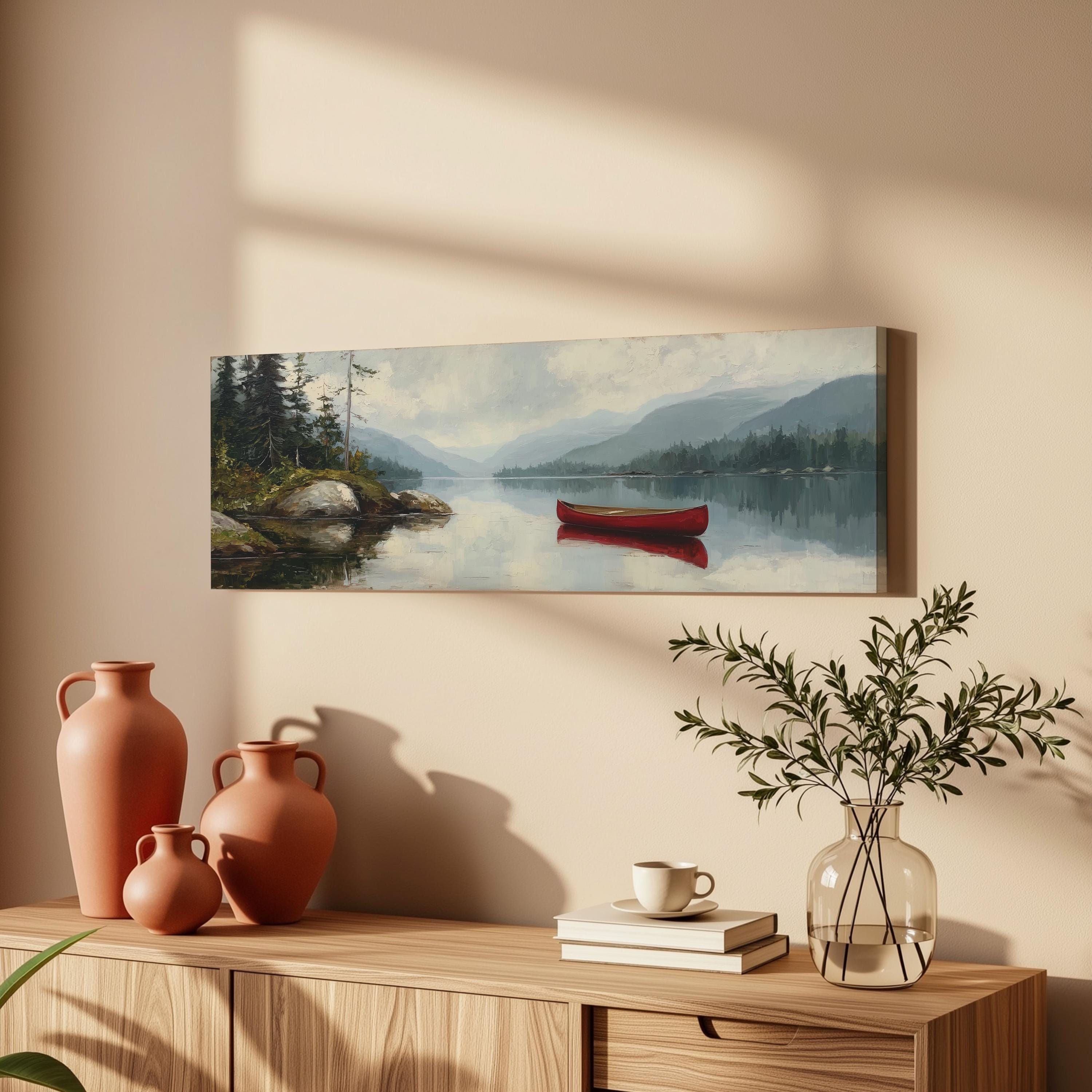 a serene landscape with a red canoe on a calm lake, surrounded by mountains and trees. The scene is captured in a painting, which is hung on a wall above a wooden cabinet. The cabinet has various decorative items, including three orange vases, a white coffee cup, and a stack of books. The overall setting creates a peaceful and inviting atmosphere.
