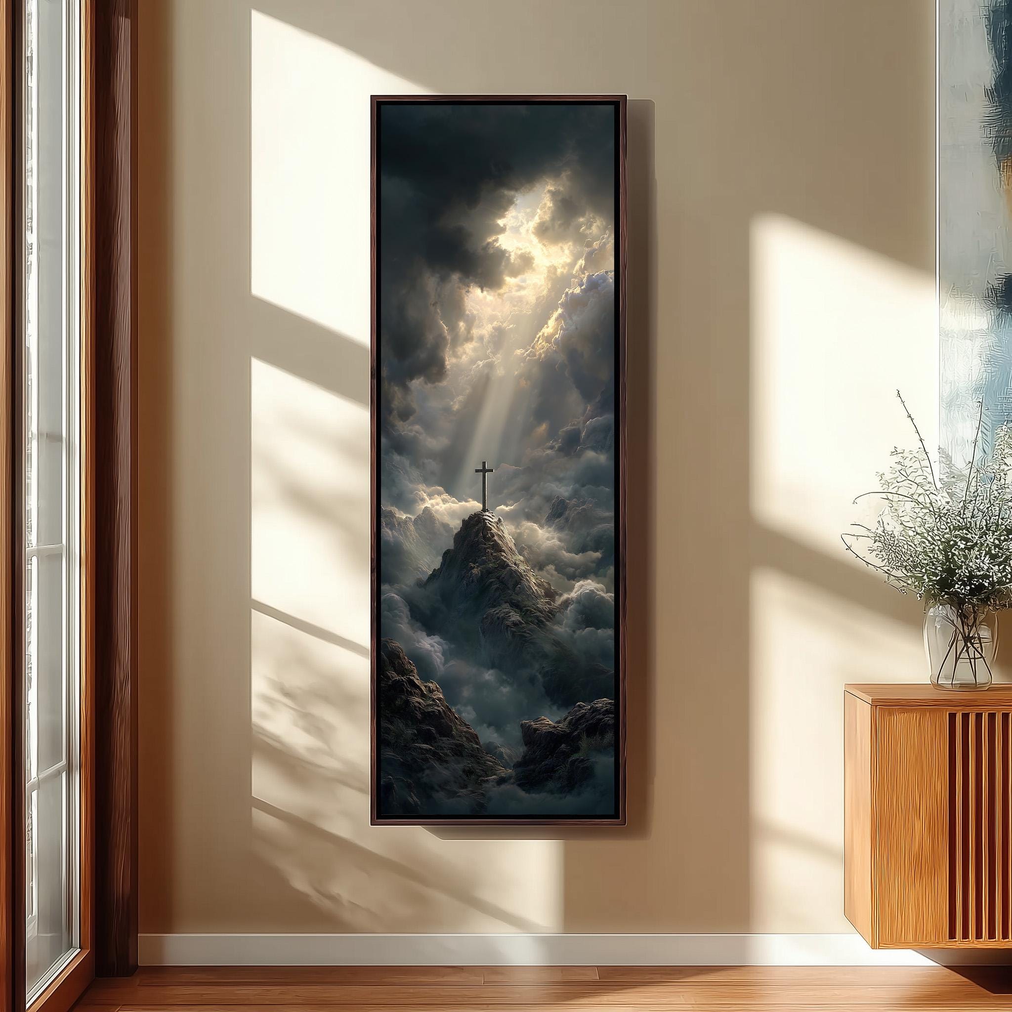 a large framed painting hanging on a wall, depicting a cross standing on a mountain peak surrounded by clouds and sunlight.
