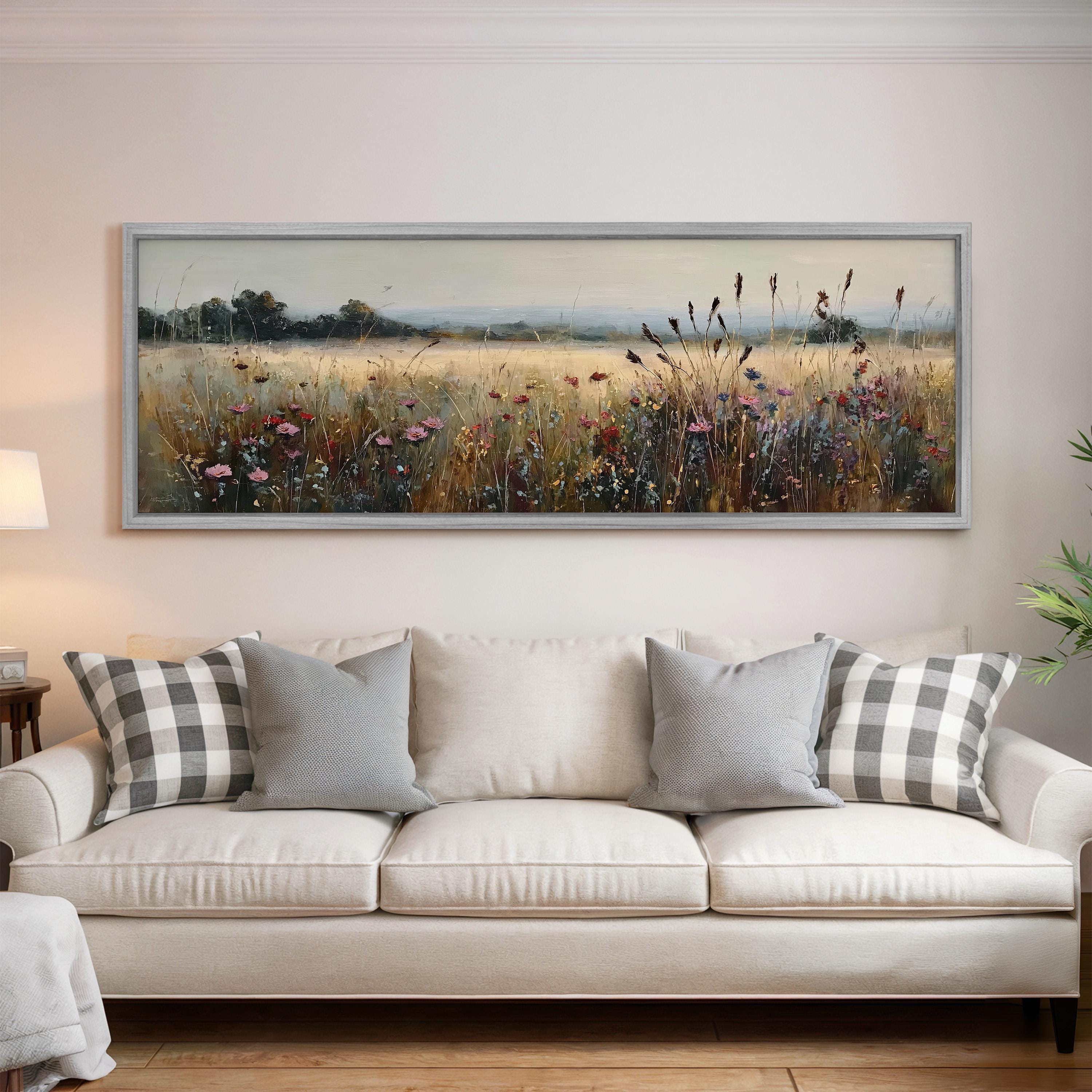 a large, framed painting of a field with wildflowers hanging on a wall above a white sofa with gray and white plaid throw pillows.