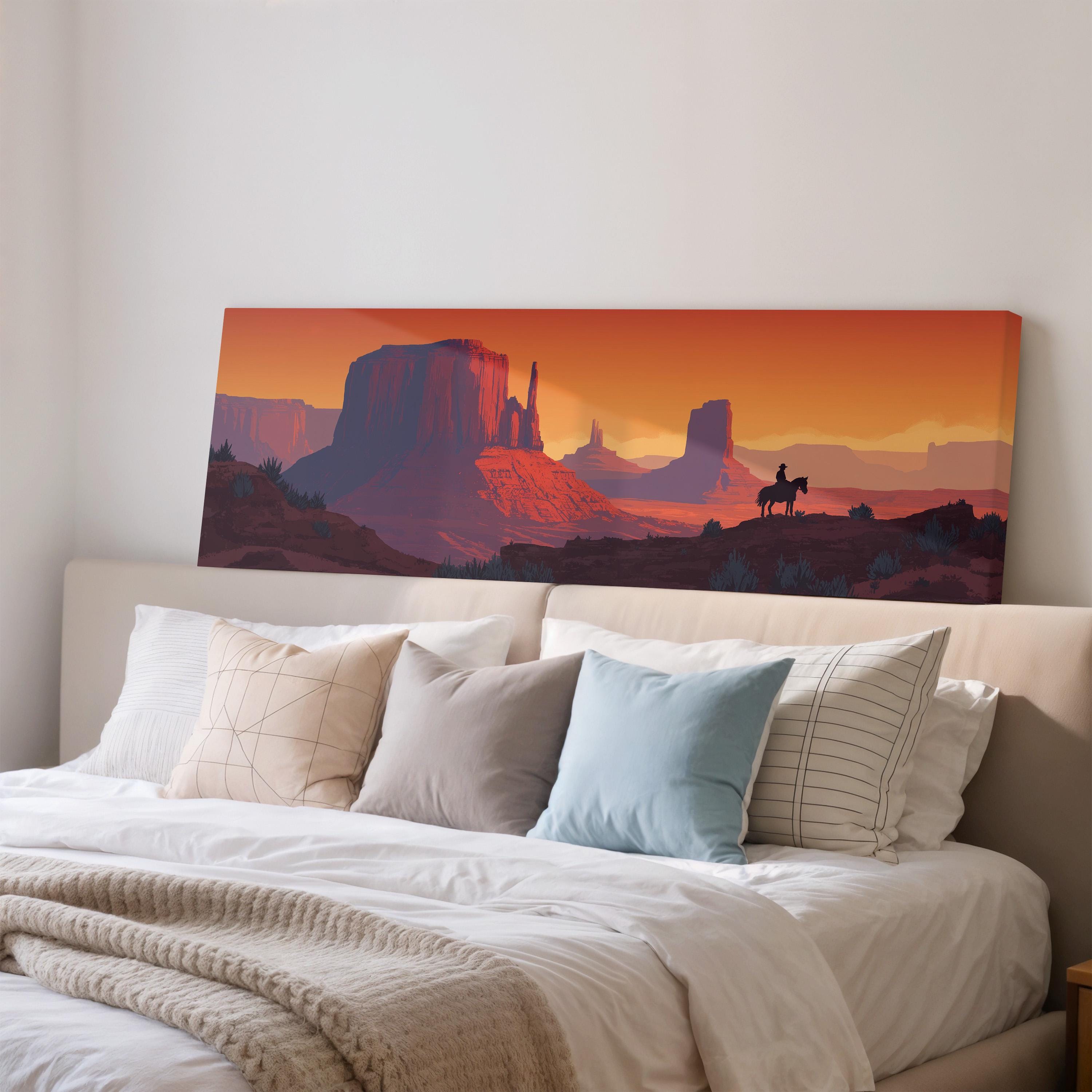 a large canvas print of a desert landscape with mountains and a cowboy on horseback, hanging on a wall above a bed with white pillows and a gray blanket.