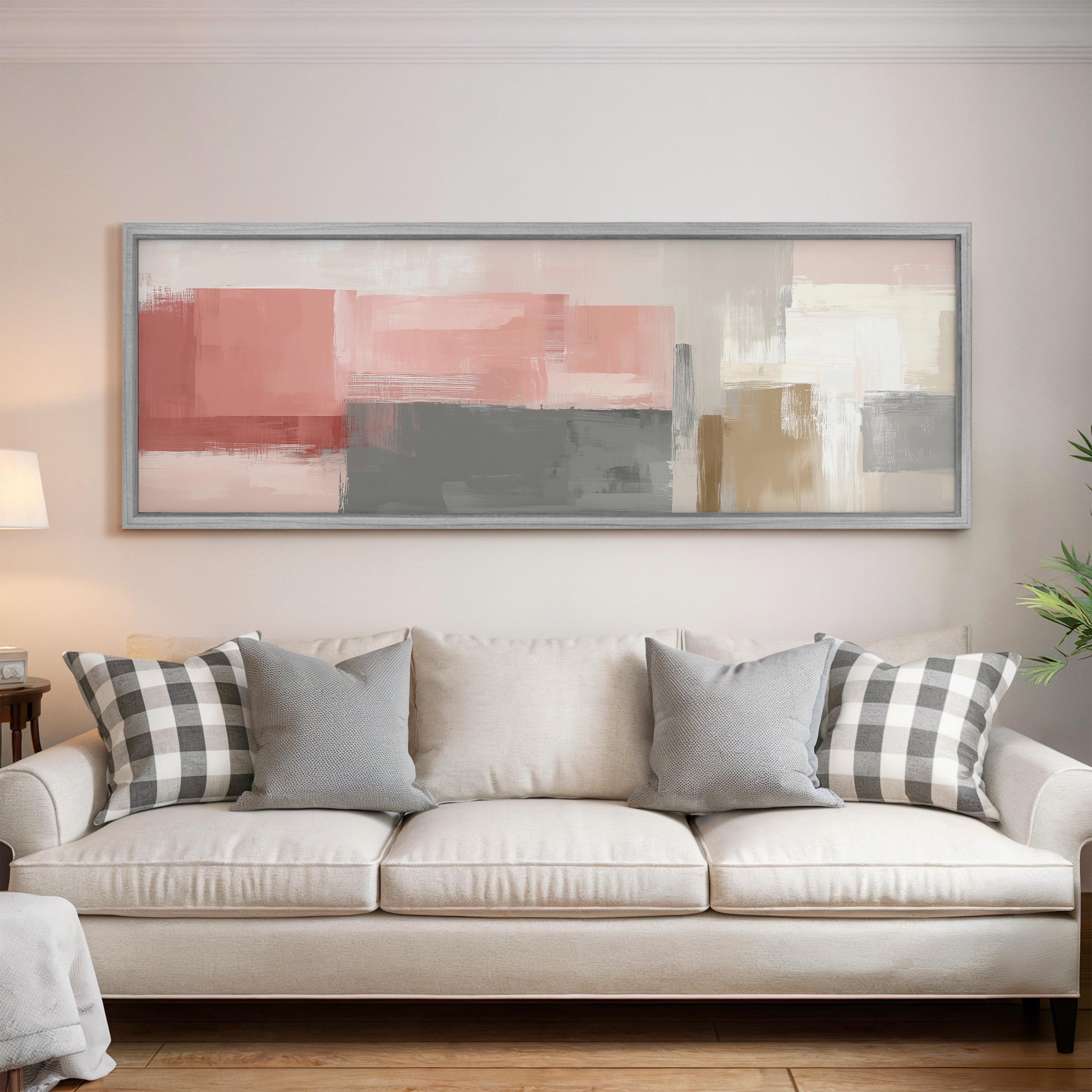 a living room with a white sofa adorned with gray and black plaid throw pillows. Above the sofa, there is a large abstract painting with a mix of pink, gray, and white colors. The room has a wooden floor and a lamp on a side table.