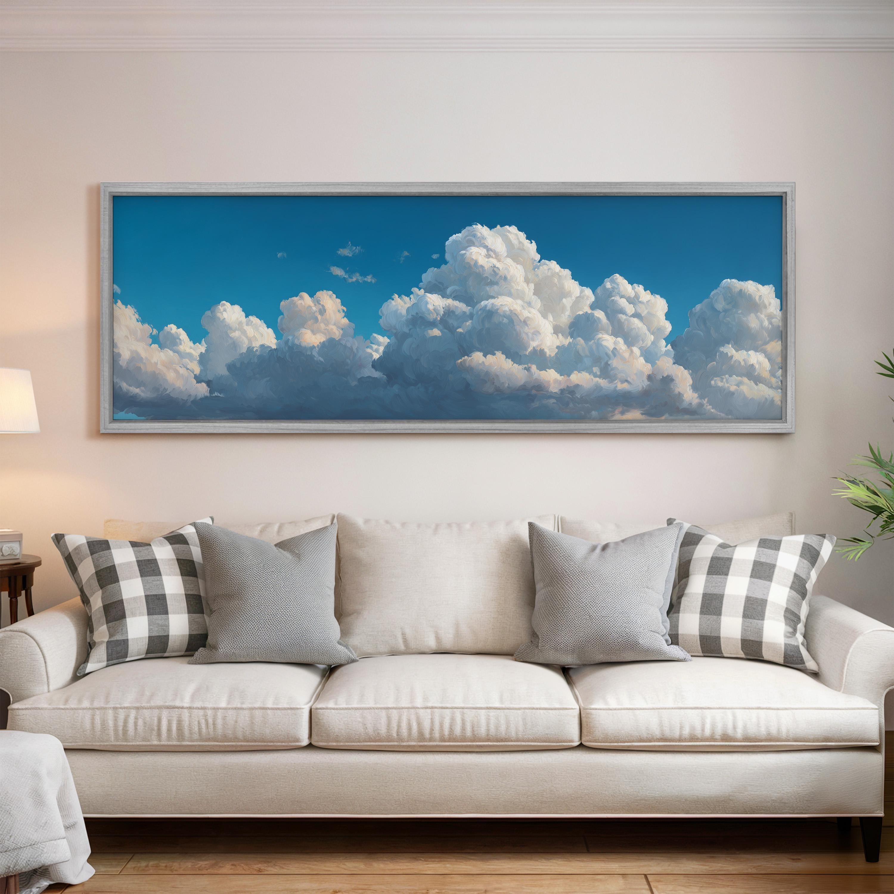 a large framed picture of a blue sky with fluffy white clouds hanging on a wall above a white couch with gray and white pillows.