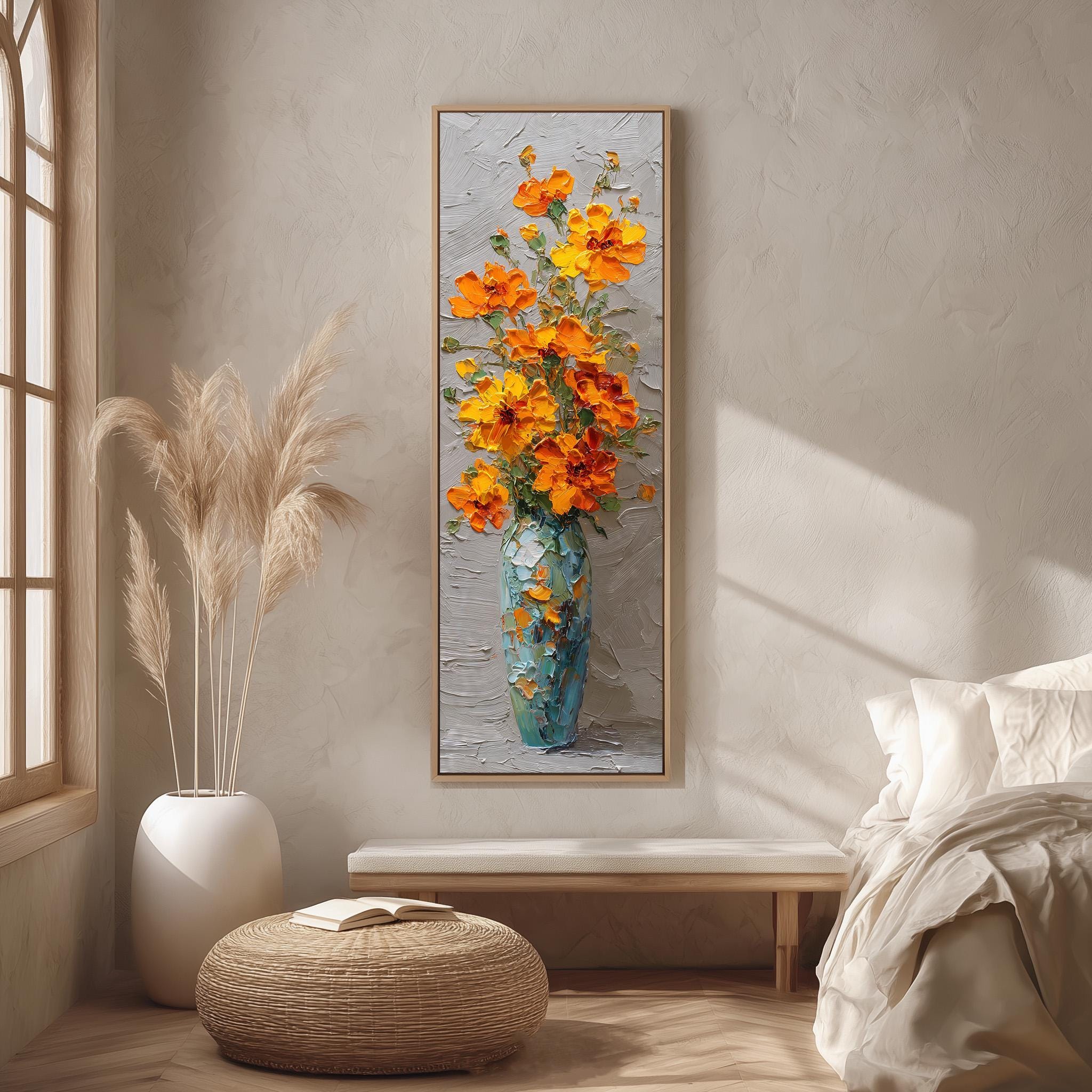 a large painting of a vase filled with orange flowers hanging on a wall in a room with a bed, a wicker ottoman, and a window.