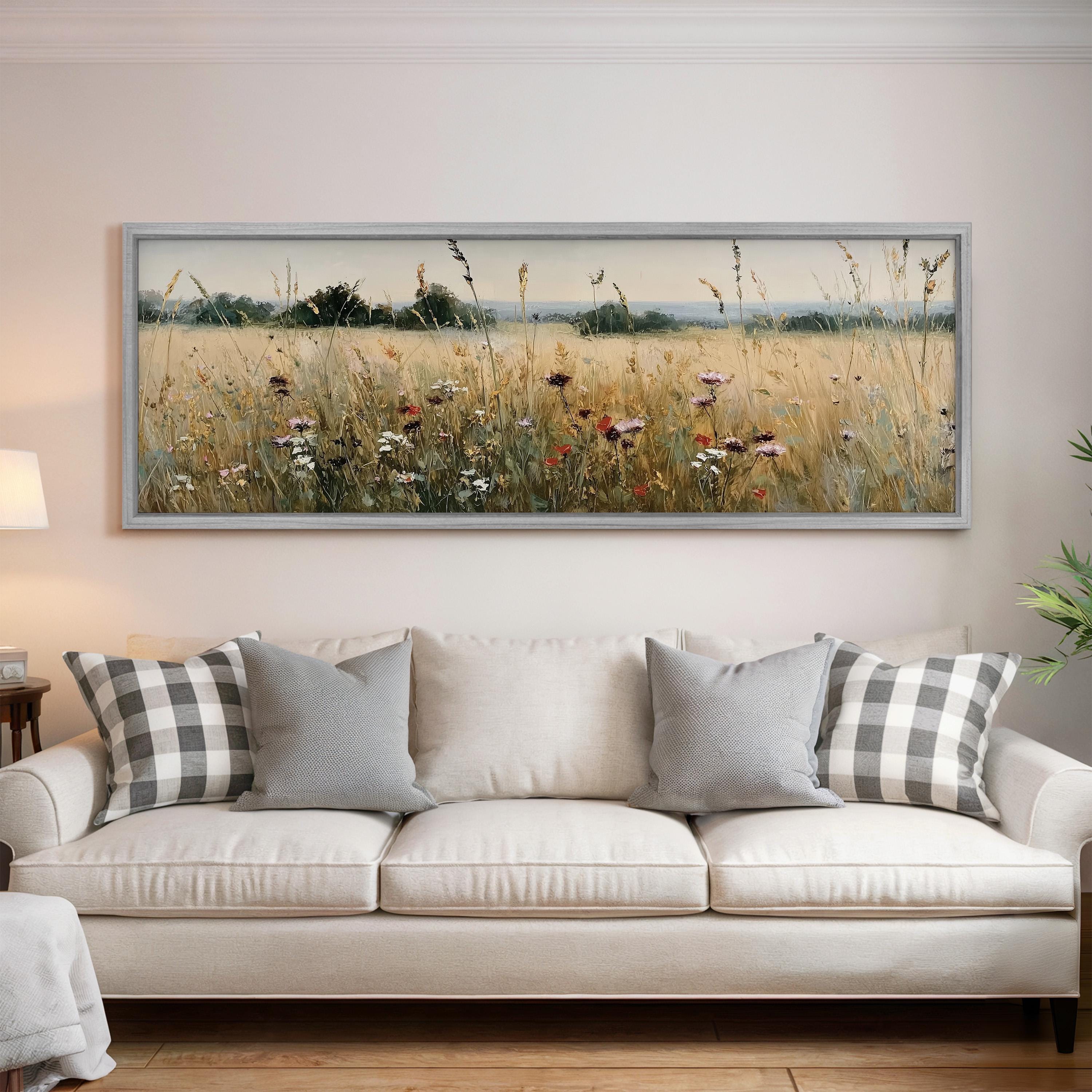a large, framed painting of a field with tall grass and wildflowers hanging on a wall above a white sofa with gray and white plaid throw pillows.