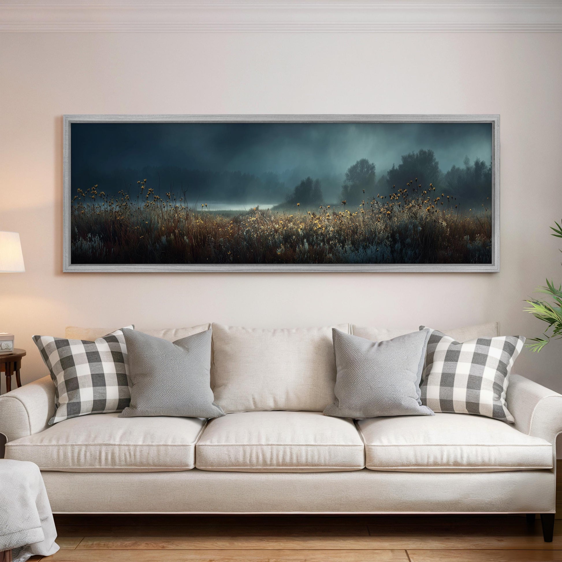 a large, framed painting of a field with tall grass and trees hanging on a wall above a white couch with gray and white plaid throw pillows.