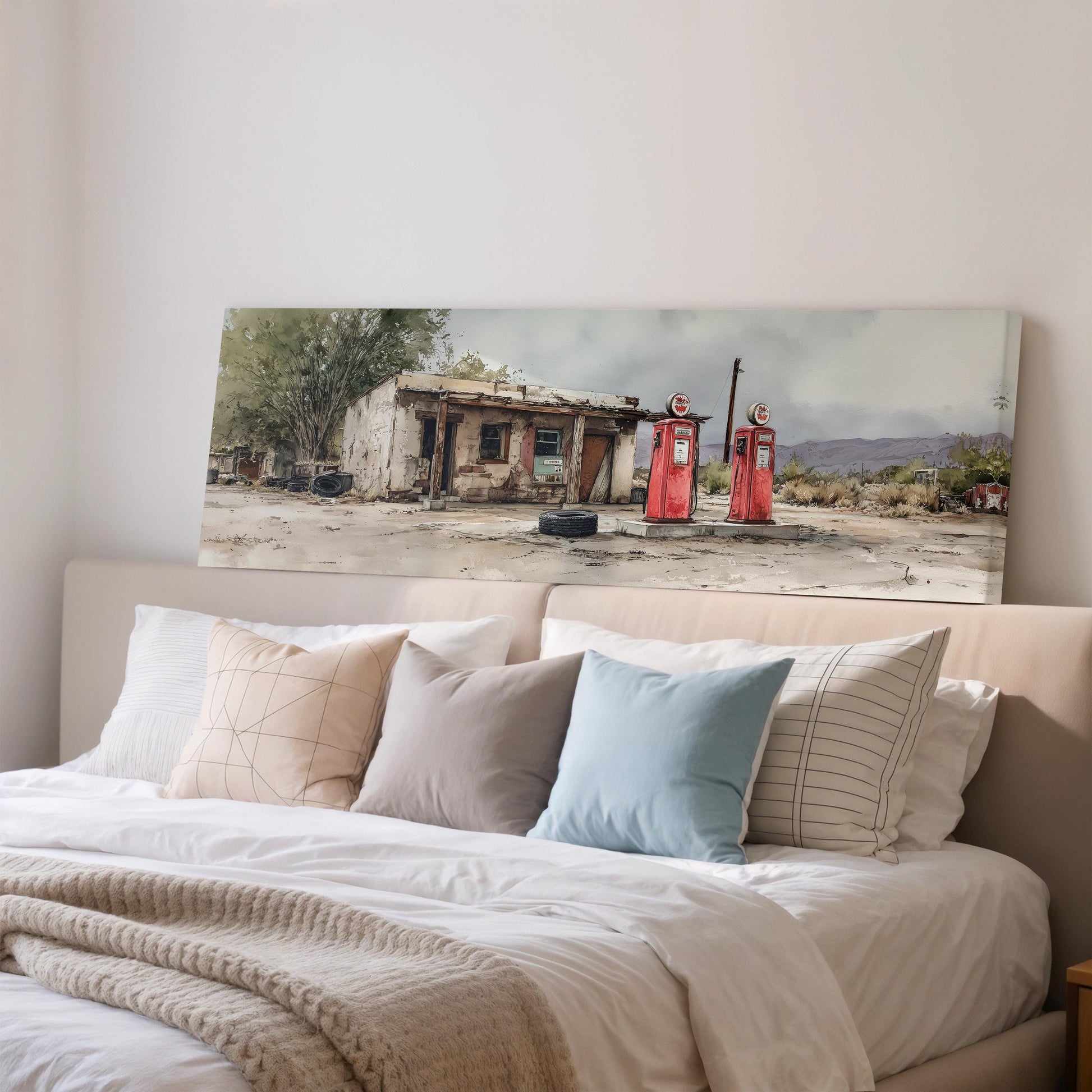 a large, white bed with a beige blanket and pillows, and a framed painting of a dilapidated building with two red gas pumps in the background.
