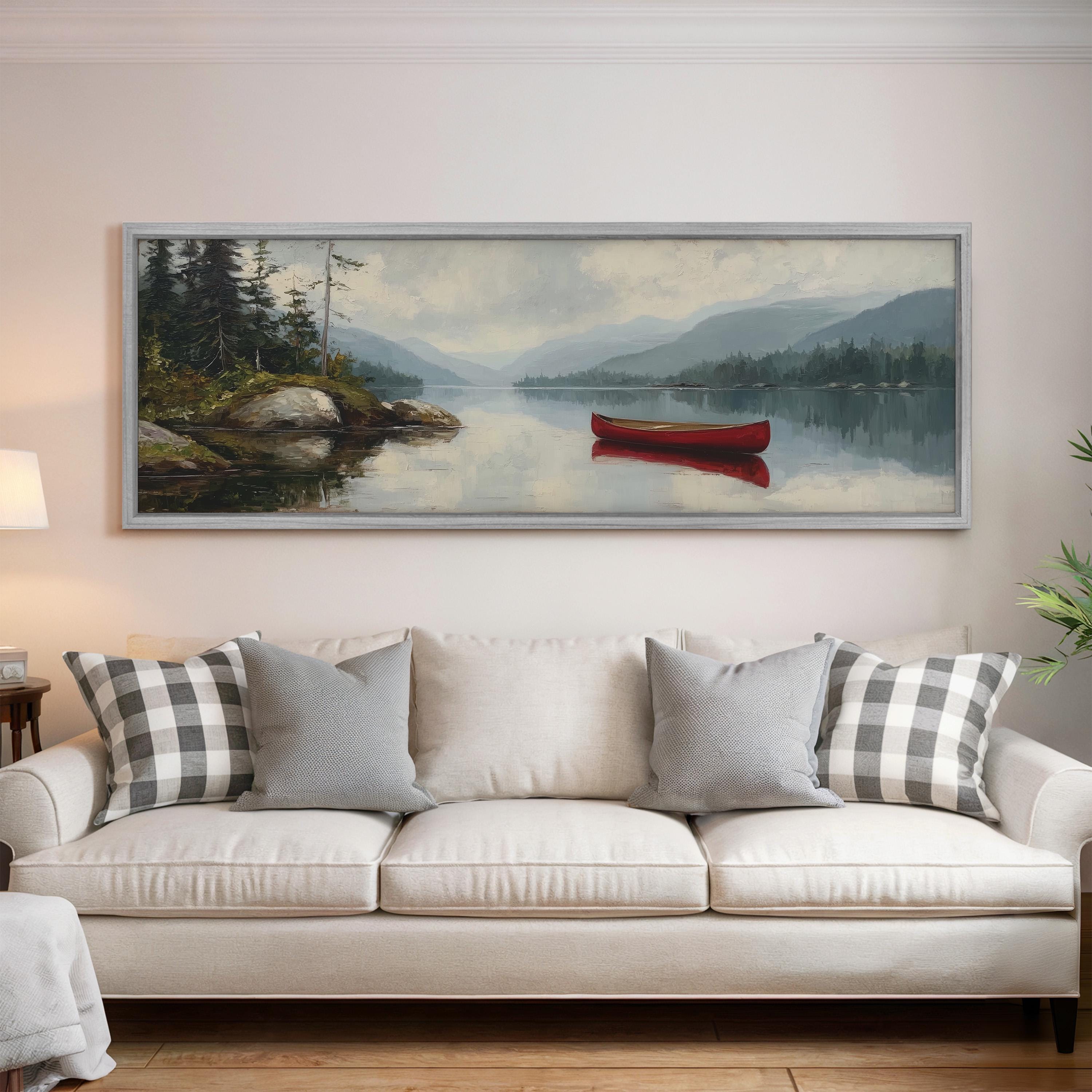 a large painting of a serene lake scene with a red canoe floating on the water, surrounded by mountains and trees. The painting is hung on a wall above a white sofa with gray and white patterned pillows.