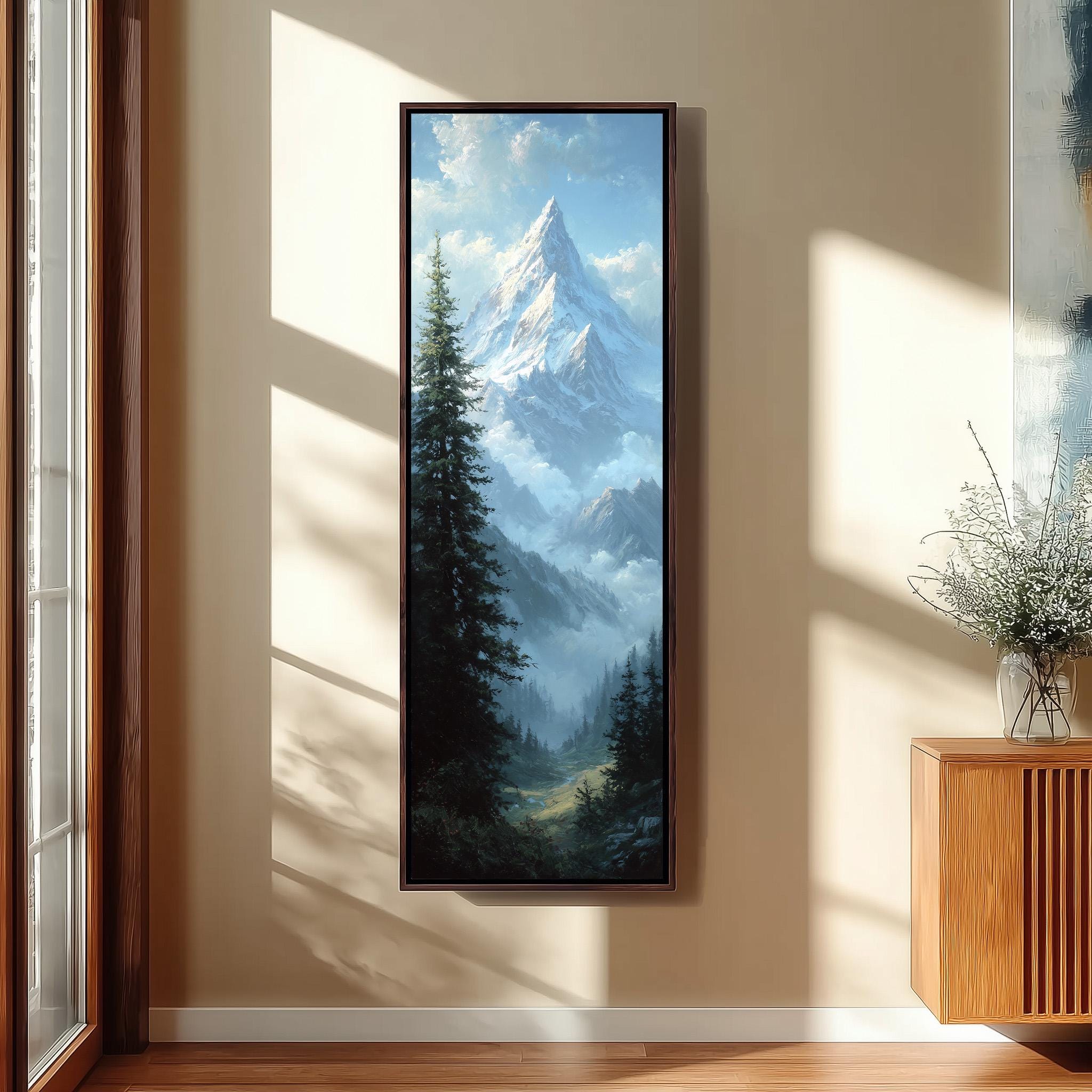 a large framed painting of a mountain landscape hanging on a wall, with a vase of flowers placed next to it.