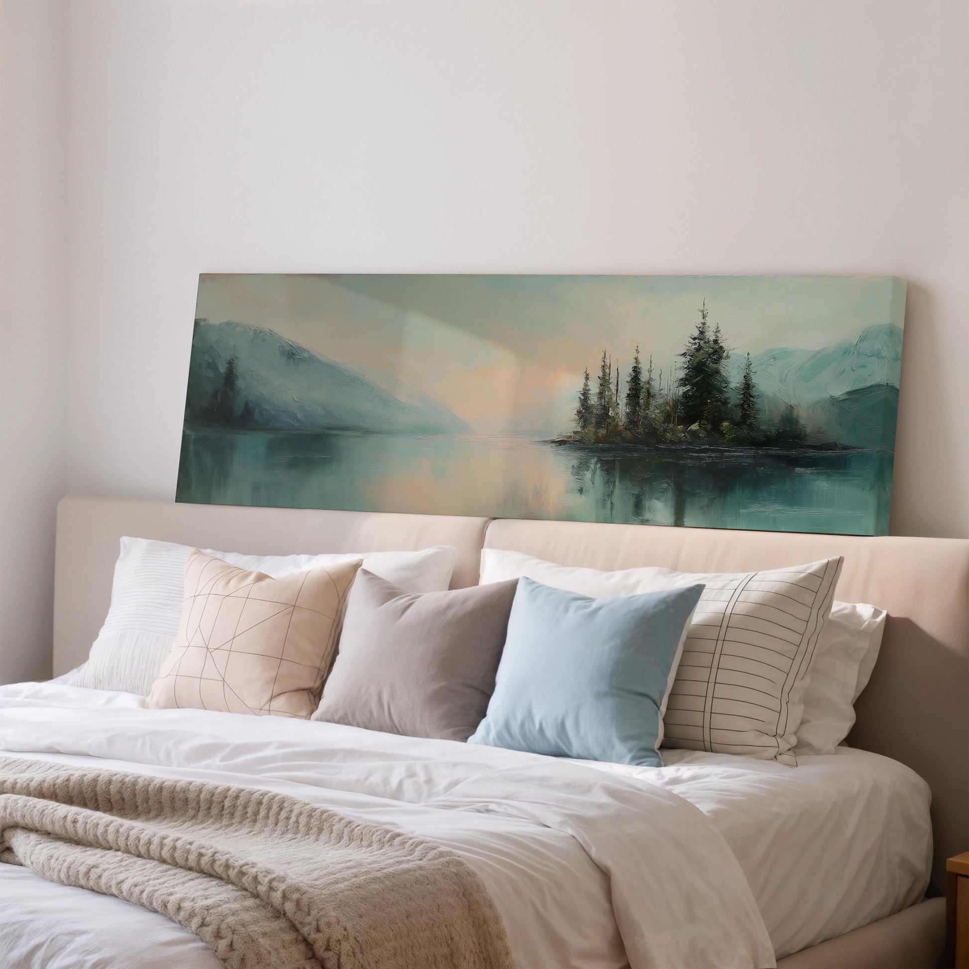 a large painting of a serene landscape with a lake and mountains in the background, hanging above a bed with white pillows and a gray blanket.