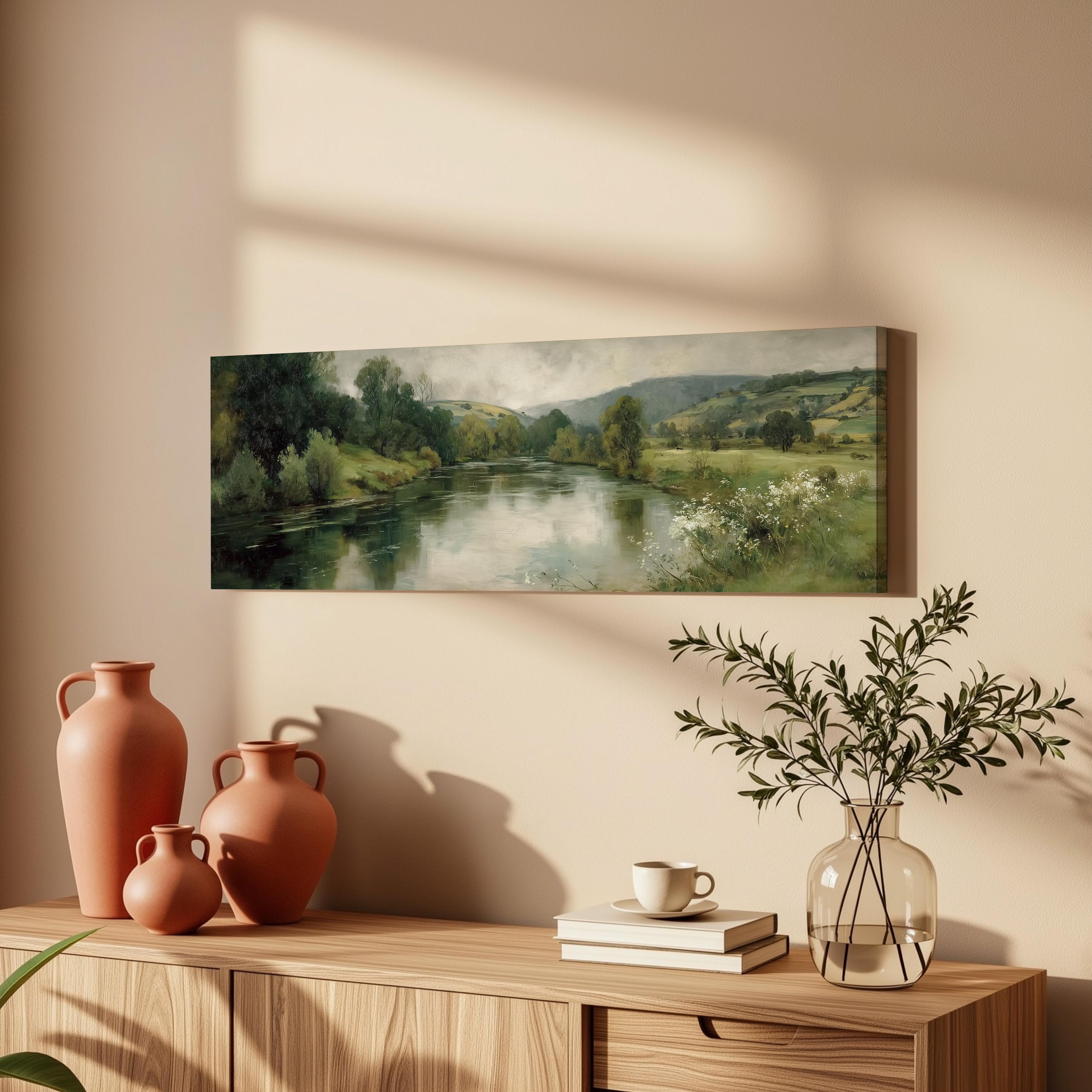 a painting of a serene landscape with a river, trees, and mountains, hanging on a wall above a wooden cabinet with various decorative items on top.