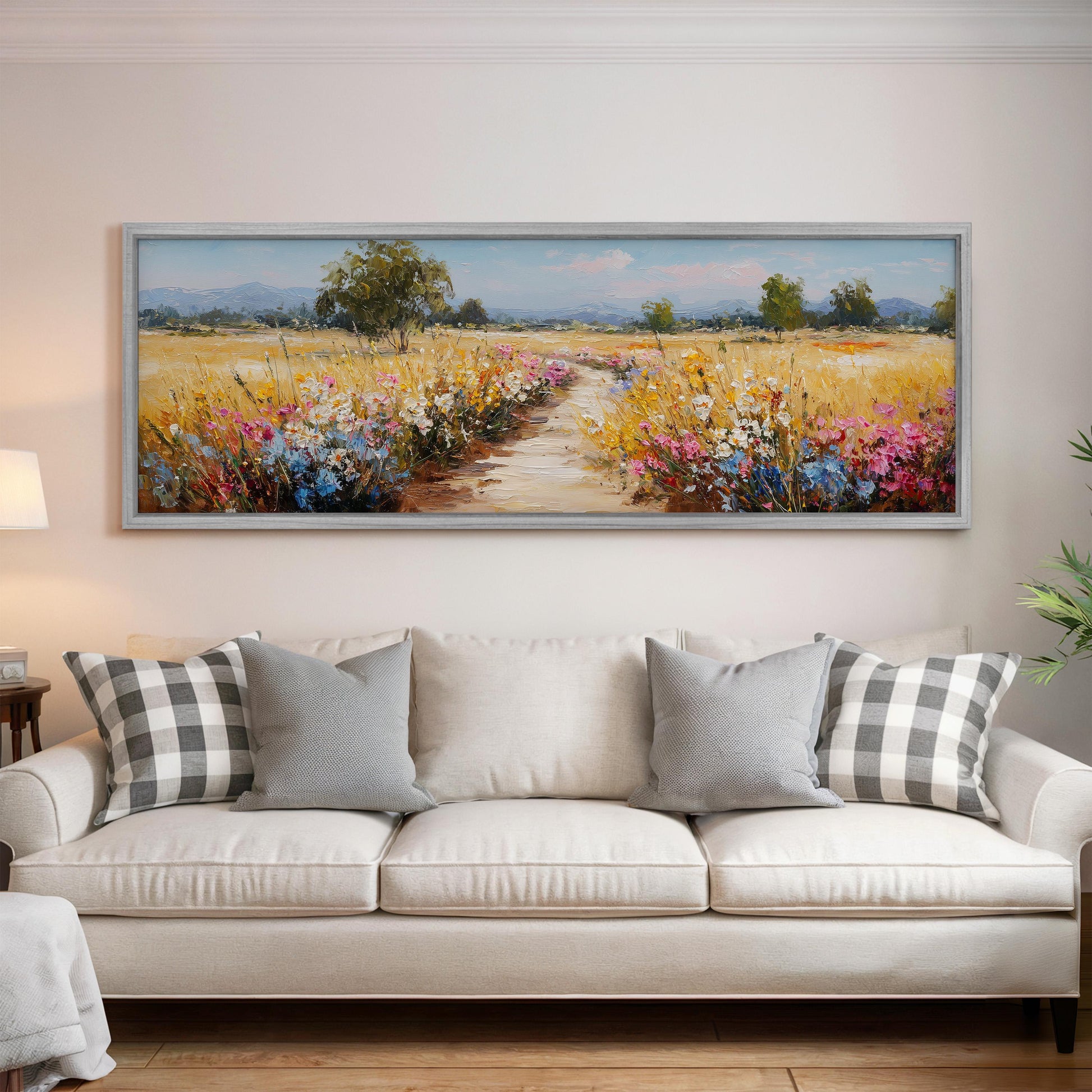 a large, framed painting of a field with flowers hanging on a wall above a white couch with gray and white plaid throw pillows.