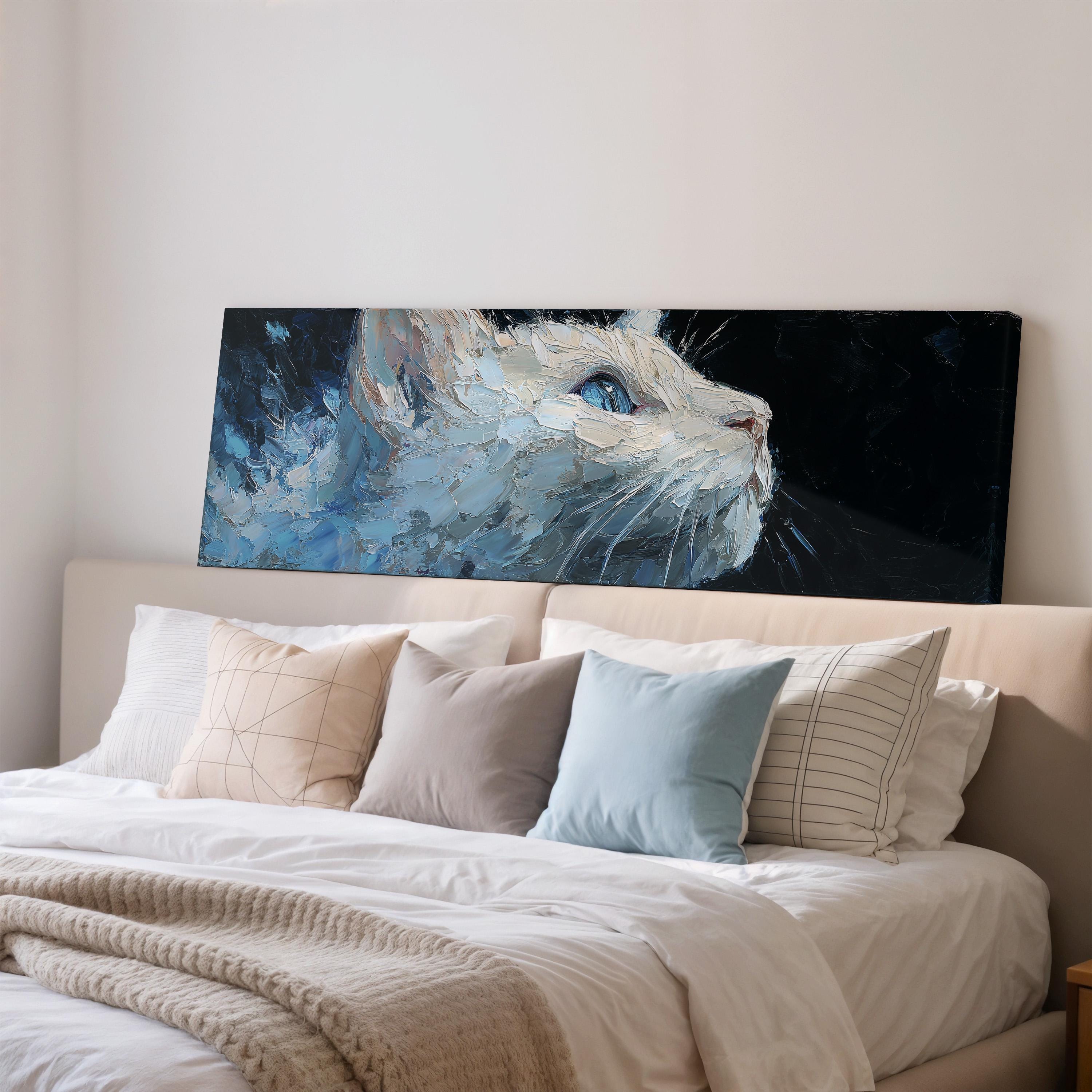 a large painting of a cat with a blue background and white fur, hanging above a bed with white pillows and a gray blanket.