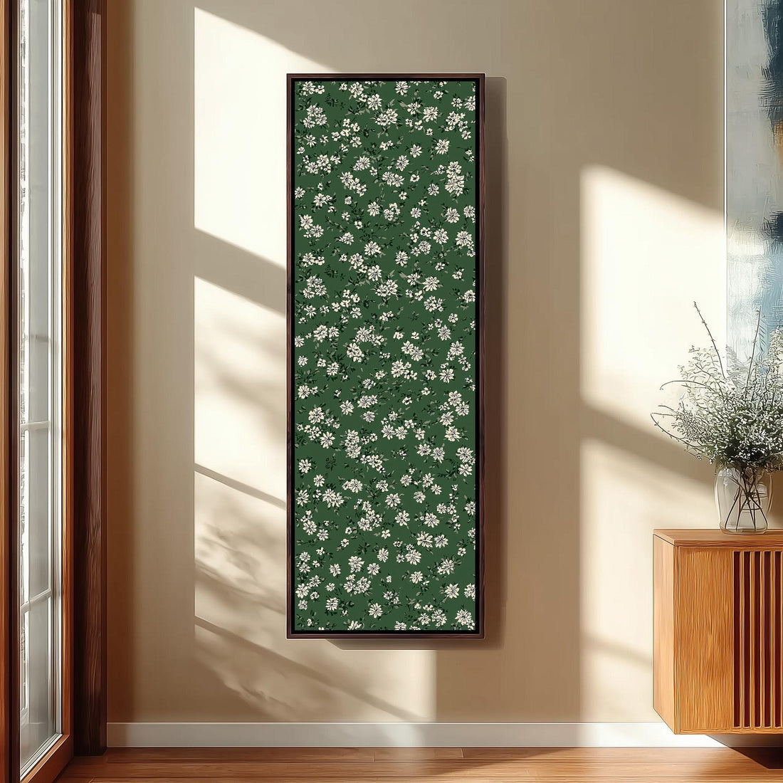 a large framed picture hanging on a wall, with a green floral pattern covering the entire frame. The picture is placed next to a wooden cabinet and a vase of flowers, creating a cozy and inviting atmosphere in the room.