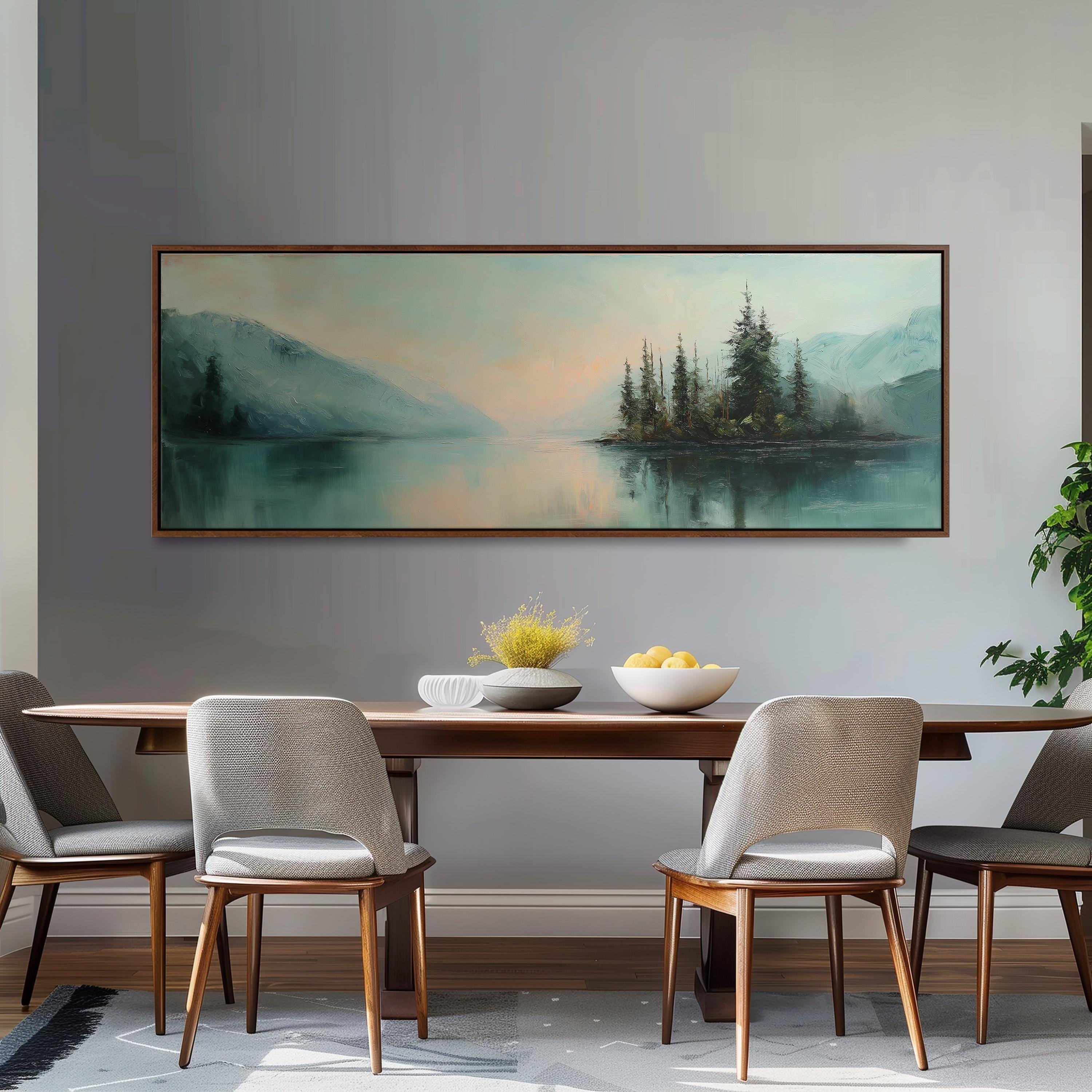 a dining room with a large painting of a serene landscape hanging on the wall above a wooden dining table with four chairs. The painting depicts a tranquil scene of a lake surrounded by mountains, with a small island in the center. The table is set with a bowl of fruit and a vase of flowers, creating a cozy and inviting atmosphere.