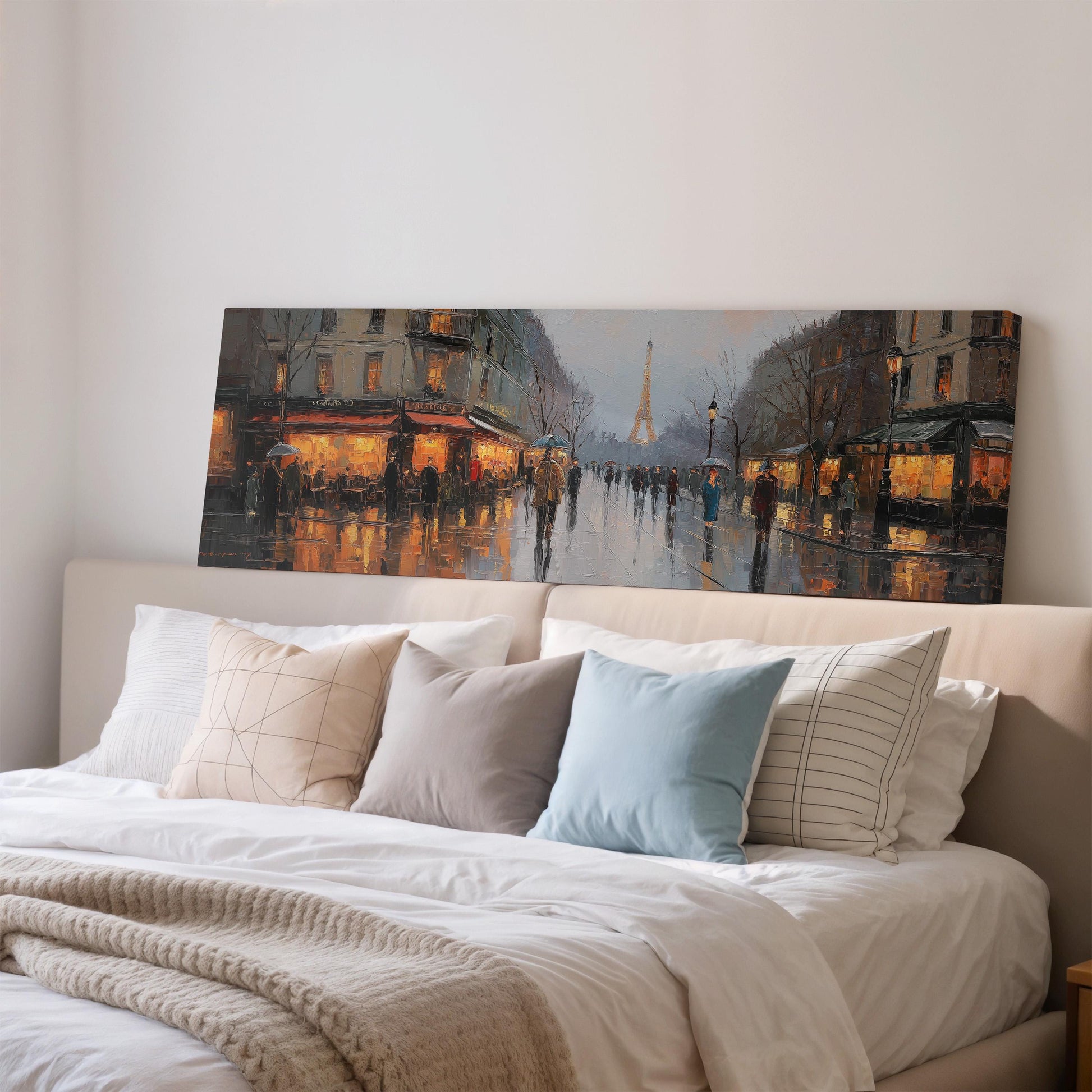 a large painting of a rainy Paris street scene hanging on a wall above a bed with white pillows and a gray blanket.