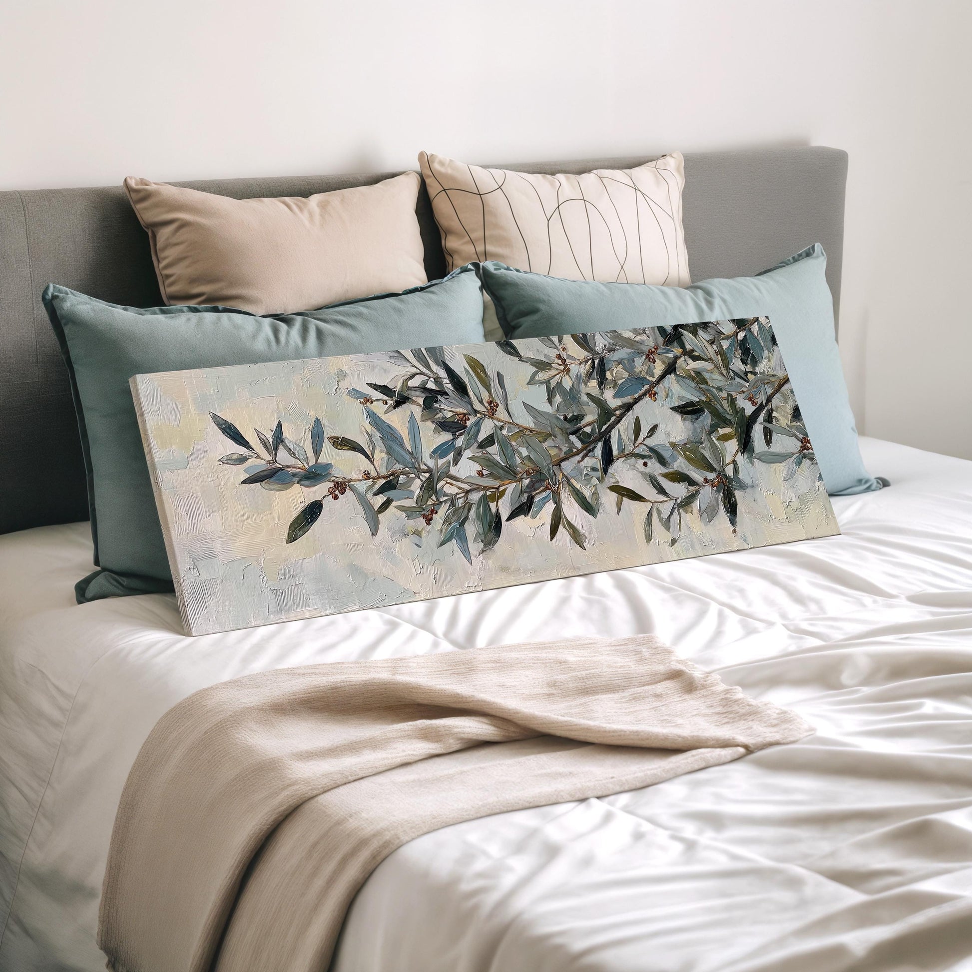 a bedroom with a bed covered in white sheets and a painting of a tree branch with green leaves and branches. The bed is decorated with multiple pillows, and there is a blanket draped over the bed.