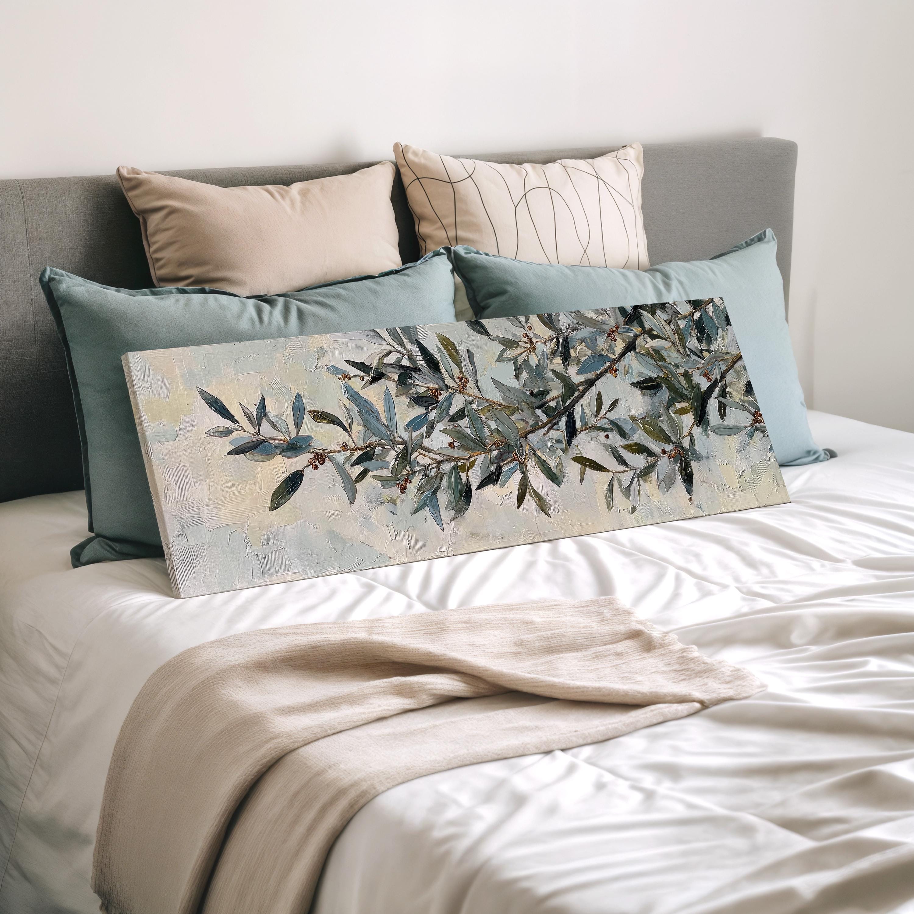 a bedroom with a bed covered in white sheets and a painting of a tree branch with green leaves and branches. The bed is decorated with multiple pillows, and there is a blanket draped over the bed.