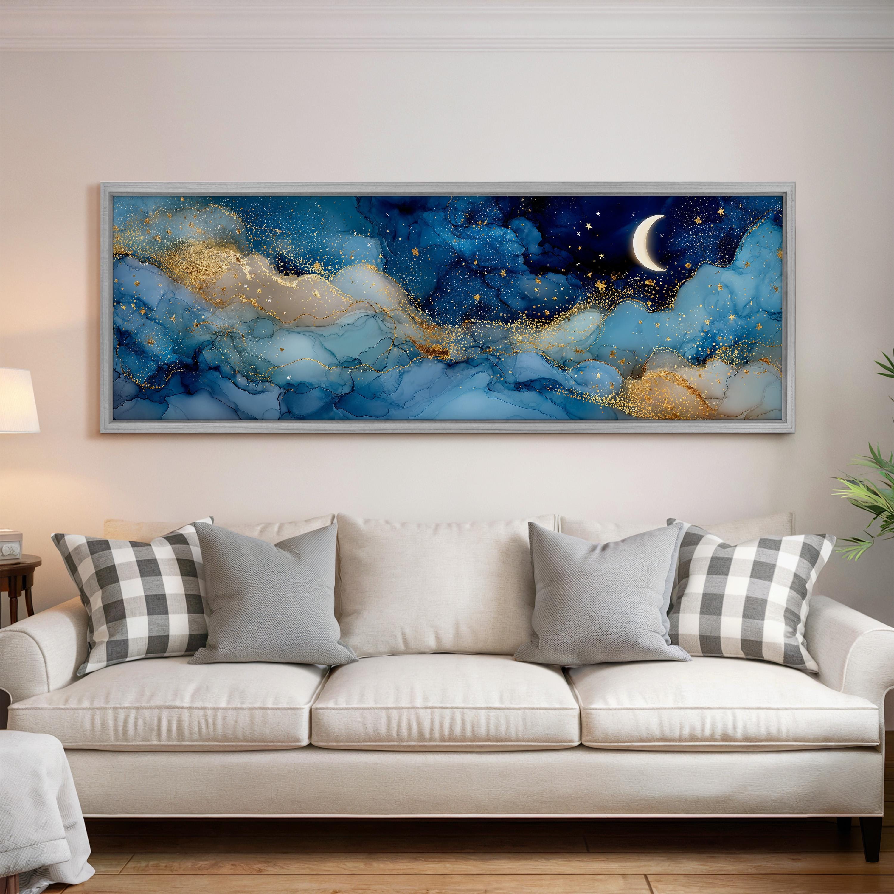 a large, framed painting of a night sky with a crescent moon and stars, hanging on a wall above a white couch with patterned throw pillows.