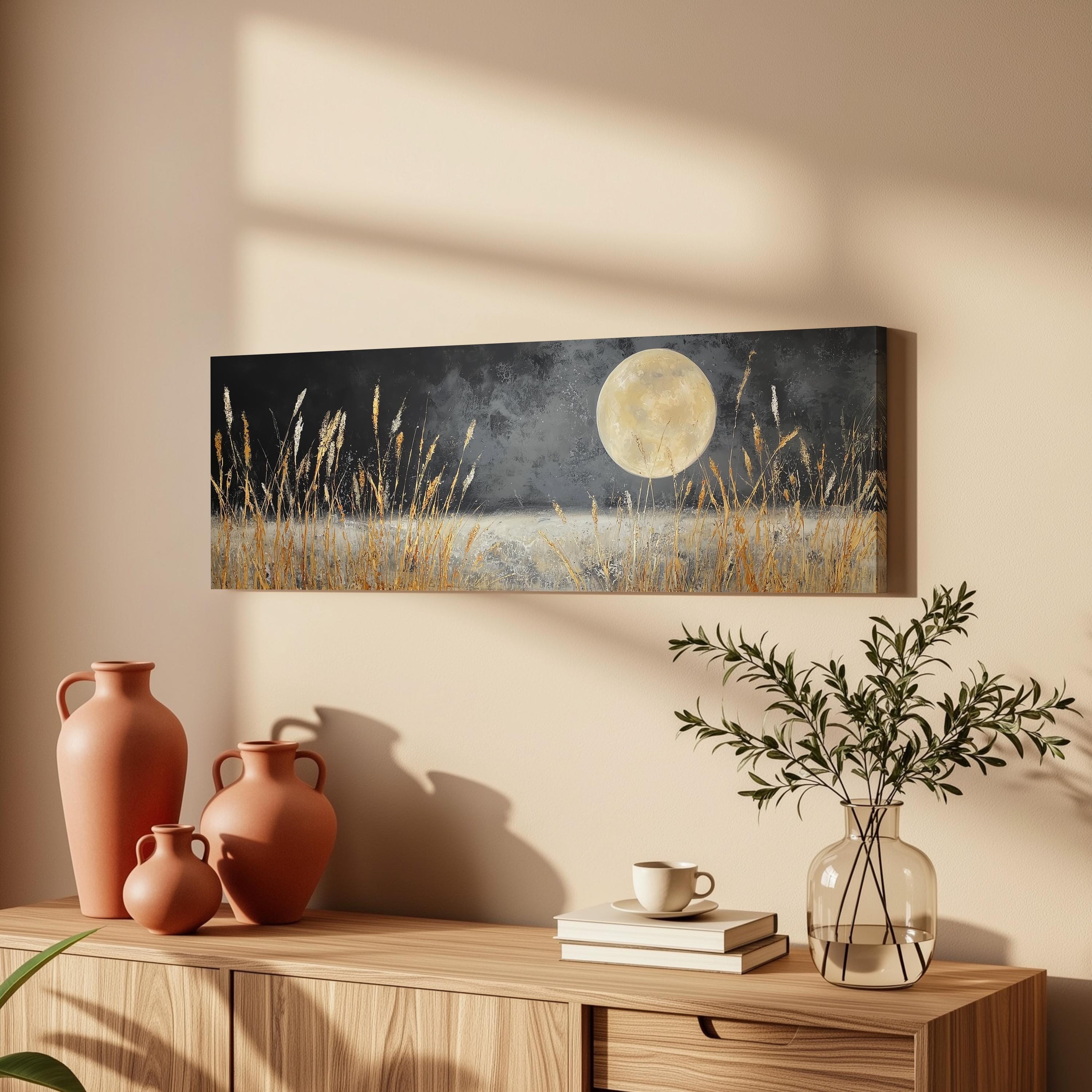 a wooden cabinet with a painting of a field of tall grass and a full moon hanging on the wall above it. The cabinet also has three orange vases and a glass vase with greenery, as well as a stack of books and a coffee cup on the surface.